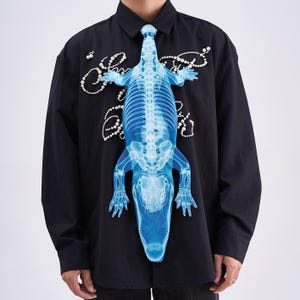 Pode incluir: Camisa preta com botões com um design de esqueleto de jacaré azul. O gráfico do jacaré é centralizado e se estende até a gola, formando uma gravata. Enfeites brancos em forma de pérola são espalhados pela camisa.