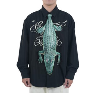 以下が含まれることがあります： 黒い長袖シャツ。緑色のワニの精巧なイラストが特徴で、ネクタイからシャツの前面にデザインが広がっています。白い筆記体のテキストと真珠のアクセントも含まれています。