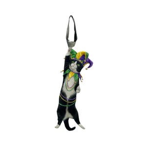 May include: A whimsical black and white cat ornament, adorned with a jester's hat in yellow, green, and purple. The cat wears a beaded necklace and has a loop for hanging. A festive decoration for Mardi Gras or any celebration.