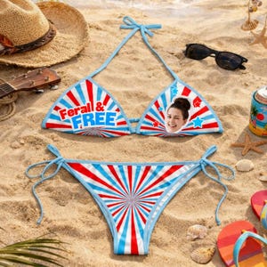 Op de afbeelding: Een rood, wit en blauw bikinipakket met de tekst "Feral & FREE" op de top. De bikinitop heeft het gezicht van een vrouw en steraccenten. De bikini ligt op een zandstrand met een zonnebril en een hoed.