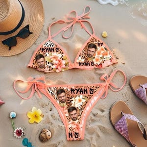 May include: A peach-colored bikini set with a leopard print design, featuring floral accents and the name "RYAN D." printed on the top and bottom. The bikini is displayed on a sandy beach with accessories.