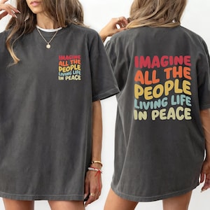 Op de afbeelding: Donkergrijs t-shirt met de tekst "IMAGINE ALL THE PEOPLE LIVING LIFE IN PEACE" in een regenboogkleurenschema op de voor- en achterkant. Het shirt heeft een relaxte pasvorm en korte mouwen.