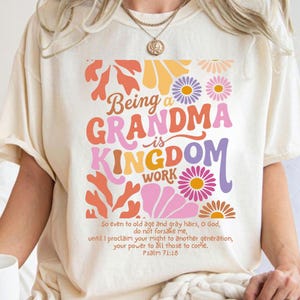 Op de afbeelding: Crèmekleurig T-shirt met een kleurrijk bloemmotief en de tekst "Being a GRANDMA is KINGDOM WORK." Het ontwerp bevat oranje, roze, gele en paarse bloemen. De onderkant van het shirt bevat een bijbelvers uit Psalm 71:18.