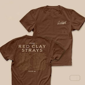 Puede incluir: Una camiseta marrón de manga corta con la palabra Grateful en letra cursiva blanca en el pecho. La parte posterior muestra el texto The Red Clay Strays y una lista de fechas y lugares de la gira Tour 26 en una fuente serif blanca.