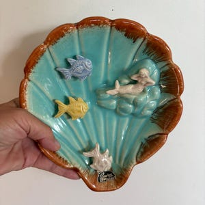 May include: A ceramic seashell-shaped dish with a turquoise interior and brown rim. The dish features three small fish and a mermaid figurine. The dish is marked with the text "Bowen Art Ware".