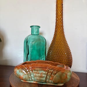 May include: A selection of vintage glass bottles and a ceramic planter. The bottles are teal and amber, with textured surfaces. The planter is oval-shaped, with a brown and teal glaze, and sits on a wooden tray.