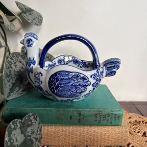 May include: A white ceramic basket shaped like a chicken with a blue handle. The body features intricate blue floral and feather patterns. The chicken head is positioned on the left side, and the tail is on the right, with a blue arched handle.