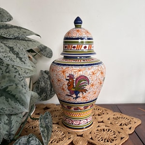 May include: A ceramic ginger jar with a lid, featuring a hand-painted rooster motif in orange, green, blue, and purple. The body is decorated with orange floral patterns and horizontal bands of colour. The lid has a pointed finial and matching stripes.