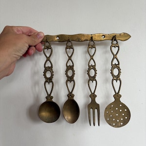May include: Vintage brass kitchen utensil set. Includes a spoon, slotted spoon, fork, and spoon. The utensils hang from a decorative brass bar. The set has an aged, antique appearance.
