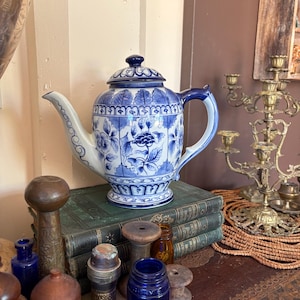 May include: A white ceramic teapot featuring a blue floral pattern, a matching lid, a curved spout, and a handle. The teapot is decorated with intricate leaf and flower motifs in a classic blue and white colour scheme, with a pedestal base.