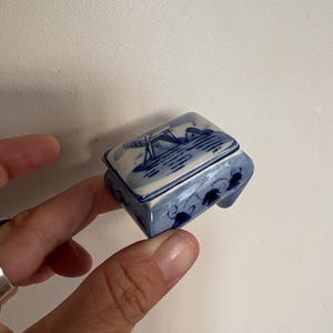 May include: Small blue and white ceramic trinket box with a lid. The box features a landscape scene on the lid and floral designs on the sides. The box is rectangular and appears to be hand-painted.