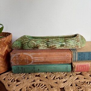 May include: A green ceramic planter with a textured, wood-like design rests on a stack of vintage books. The books have aged covers in shades of orange, green, and blue. A woven basket is visible on the left.