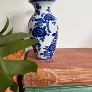 May include: A white vase with a blue floral pattern. The vase has a flared rim and a narrow neck. The blue floral pattern is a classic design. The vase is on top of a stack of vintage books.