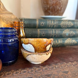 May include: Two small, round ceramic bowls stacked vertically. The bowls feature a textured, mottled glaze with a combination of earthy brown and off-white tones. The glaze appears to drip down the sides, creating a layered, organic appearance.