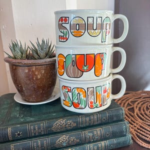 May include: A stack of three off-white ceramic soup mugs, each featuring the word SOUP in large, stylised letters filled with colourful illustrations of various vegetables, including carrots, tomatoes, onions, and corn, with a handle on the right side.