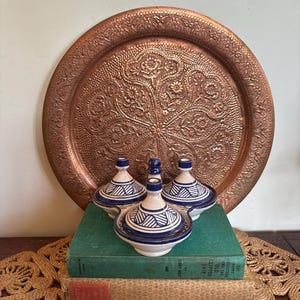 May include: Three small, white and blue ceramic tagine pots with geometric designs sit on a stack of books in front of a large, round, copper tray with an embossed floral pattern. The tray is a warm, reddish-brown colour.
