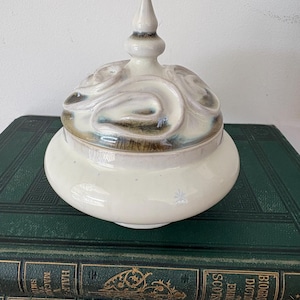 May include: A cream-coloured ceramic jar with a lid, featuring a swirling design in shades of brown and blue. The jar has a rounded shape and a pointed finial on top. The jar is sitting on top of a stack of green books.