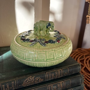 May include: A round, light green ceramic lidded jar featuring a moulded elephant figurine on top. The lid is decorated with a raised garland of dark berries and blue ribbons, while the base displays a textured grid pattern across its surface.