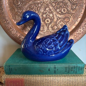 May include: A glossy, cobalt blue ceramic swan ornament with textured detailing. It rests on a stack of vintage books. The background features a copper-toned tray with an embossed floral design.