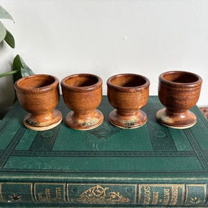 May include: Four brown ceramic egg cups with a speckled finish. The egg cups have a rounded bowl shape and a pedestal base. They are arranged on a green book.