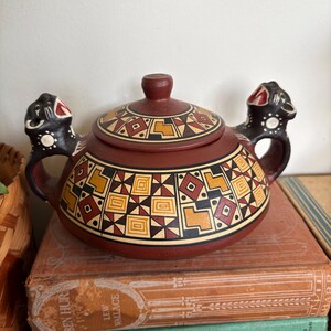 May include: A maroon ceramic teapot with a lid, featuring a geometric pattern in yellow, black, and red. The teapot has two handles with black jaguar-like figures. The teapot is sitting on top of a stack of old books.