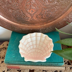 May include: A small, pale pink seashell-shaped dish with a textured surface. The dish is on a stack of vintage books with teal covers. A large, ornate copper tray is in the background.