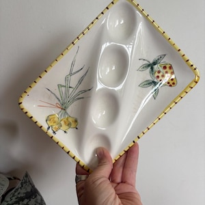 May include: White ceramic egg serving dish with a yellow dotted border. The dish has four egg indentations and is decorated with hand-painted illustrations of onions and a strawberry. The dish is held by a person's hand.