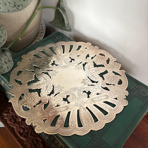 May include: A silver-coloured, ornate metal trivet with a floral and leaf design. The trivet is round with a detailed, openwork pattern. It is resting on a dark green book, suggesting a vintage or antique aesthetic. The design includes flowers and leaves.