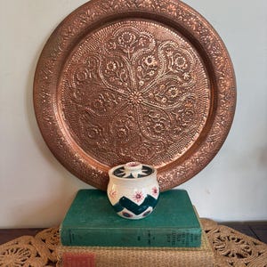 May include: A large, round copper tray with detailed floral and geometric designs. A small, white ceramic jar with a lid, adorned with pink flowers and green zigzags, rests on two vintage books. The tray and jar are displayed on a woven mat.