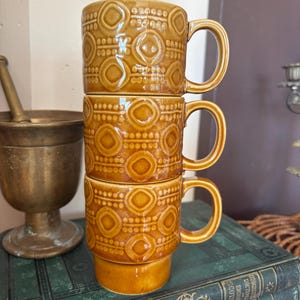 May include: A stack of three matching mustard-yellow ceramic mugs, each featuring a raised geometric pattern of circles and dots. The mugs have a glossy finish and a cylindrical shape with a side handle, designed to stack securely on top of one another.