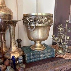 May include: A brass wine cooler or planter featuring a wide, flared rim with a relief pattern of roses and leaves. The vessel has a pedestal base and two decorative ring handles on opposite sides, resting atop a stack of three green books.
