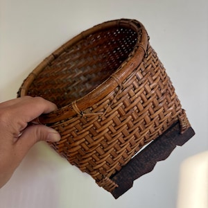 May include: A round, woven brown wicker basket featuring a thick, reinforced circular rim and a dark, scalloped wooden base. The basket is constructed with a tight, interlocking weave pattern and exhibits a natural, deep brown colour throughout.
