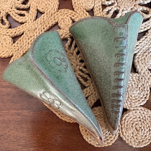 May include: Two green ceramic wall vases with a textured finish. One vase has a floral design, the other has a vertical line design. The vases are displayed on a woven mat and wooden surface.