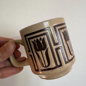 May include: A beige ceramic mug with a speckled finish and a dark brown geometric pattern featuring stylised tulip motifs. The mug has a cylindrical body with a stepped base and a sturdy handle on the side, showcasing a repeating floral design.