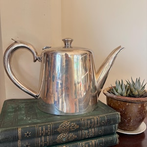 May include: A polished silver teapot with a curved handle, a straight spout, and a small lid knob. The teapot has a reflective metallic surface and sits atop a stack of three dark green hardcover books with gold embossed lettering on the spines.