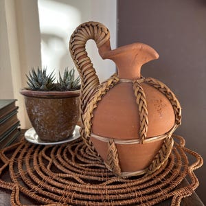 May include: A terracotta pitcher featuring a rounded body and a flared spout, encased in a decorative woven wicker cage that extends to form a sturdy handle. The natural brown clay contrasts with the lighter tan woven reed accents.