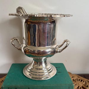 May include: A silver wine cooler with a decorative scalloped edge and two handles. The cooler is set on a pedestal base. It is displayed on a stack of books with a green cover.