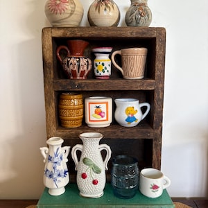 May include: A wooden shelf displays a collection of vintage ceramic vases and mugs. The vases feature floral and abstract designs, while the mugs showcase various patterns and illustrations. The shelf is placed on a stack of books.
