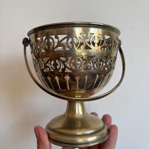 May include: A brass-coloured metal bowl with a pedestal base and a swing handle. The upper portion of the bowl features an intricate, openwork cutout design with geometric patterns, while the lower section has vertical etched lines.