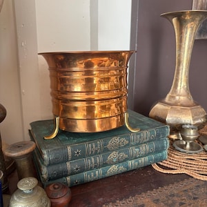 May include: A polished copper planter with a wide, flared rim and horizontal ribbed detailing, supported by three curved brass legs. The planter is resting on a stack of three green, cloth-bound books with gold-embossed lettering on the spines.