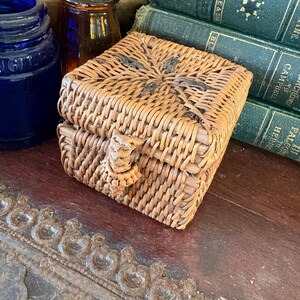 May include: A small square woven wicker box with a hinged lid and a central braided closure. The natural tan material features a star-like pattern woven into the top surface. The box is positioned near a blue glass bottle and green books.