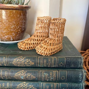 May include: A pair of small, light brown woven wicker boots with an open lattice design. The boots are crafted from thin, flexible strips of natural material, featuring a rounded toe and a tall, straight shaft that mimics the shape of a standard boot.