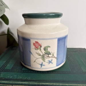 May include: A ceramic jar with a cream body, blue vertical stripes, and a green rim. The jar features a floral design with a red rose and blue flowers. The jar is on a stack of green books.