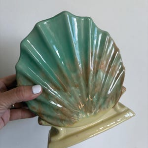 May include: A ceramic shell-shaped decorative object featuring a glossy finish with a gradient of mint green and brown hues on the fan-shaped top, supported by a solid pale yellow rectangular base with a smooth, curved design.