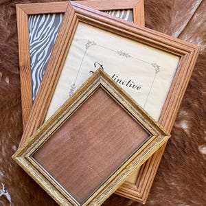 May include: Three rectangular picture frames stacked together. The largest frame has a light wood finish and contains a zebra print. The middle frame is light wood with a cream insert featuring the text "Distinctive". The smallest frame is gold.