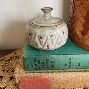 May include: A small, round, speckled ceramic jar with a lid. The jar has a textured, patterned design and sits on a stack of vintage books. A woven basket is in the background.