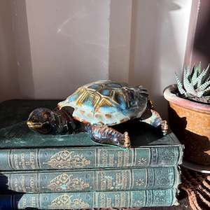 May include: A ceramic turtle figurine with a glossy, multi-coloured shell featuring shades of blue, brown, and cream. The turtle has a dark, mottled head and limbs, and it is positioned resting on top of a stack of three antique green hardcover books.