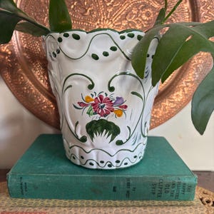 May include: A white ceramic planter with a scalloped rim and green painted trim. The body features a hand-painted floral motif in red, yellow, and green. The planter sits atop a green book titled The Happy Hollisters and the Old Clipper Ship.