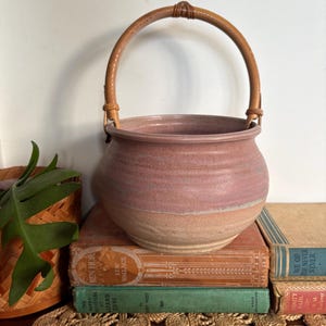 May include: A handmade ceramic pot with a light brown base and a pinkish-purple upper section. It has a woven, light brown handle. The pot is displayed on a stack of vintage books with a woven placemat.