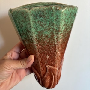 May include: A triangular wall pocket vase with a two-tone glaze. The top is a speckled green, transitioning to a reddish-brown at the bottom. The base features a stylized floral design.